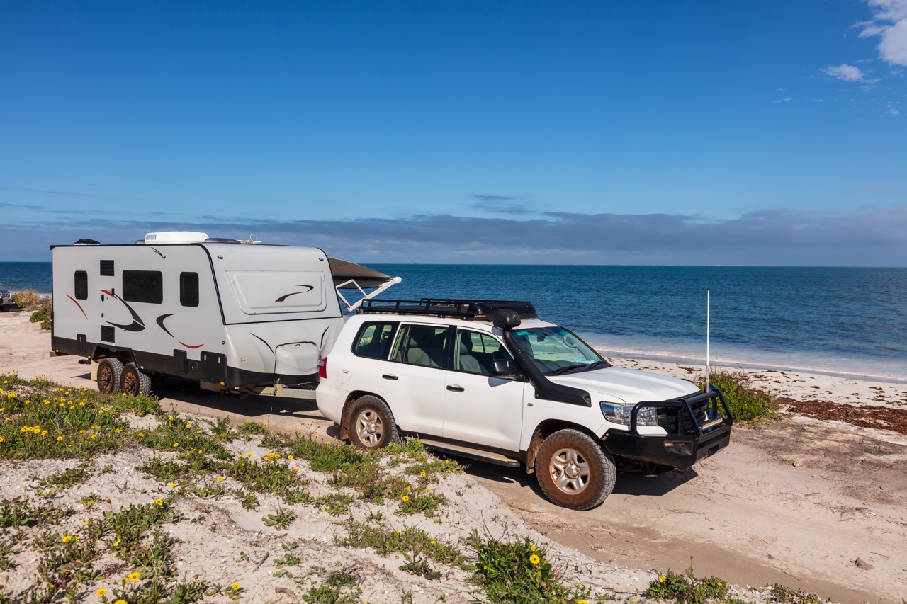 Landscape aerial view of 4WD accessories and 4WD and modern caravan parked adjacent to a sparkling sunny beach.